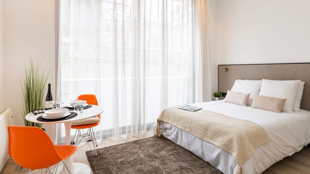 Une chambre d’hôtel moderne avec un grand lit, une table à manger avec des chaises et une grande fenêtre agrémentée de rideaux légers.