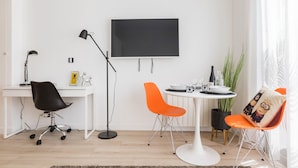 Un salon moderne avec un bureau blanc, des chaises noires et orange, une table ronde, un téléviseur à écran plat et une plante en pot.