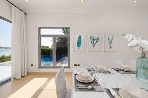 A dining area with a view of a swimming pool, a large window, and three framed botanical prints on the wall.