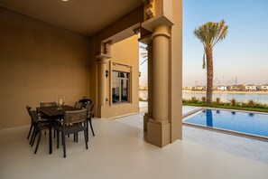 A dining area with a table and chairs, a view of a swimming pool, and a palm tree. - Exclusive Villa w/ Private Pool, Garden & Beach (Dubai)