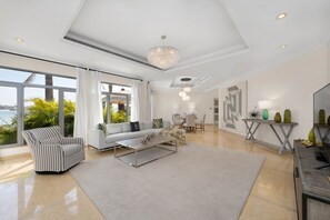 A spacious living room with a large window, a chandelier, a sofa, and a coffee table. - Exclusive Villa w/ Private Pool, Garden & Beach (Dubai)