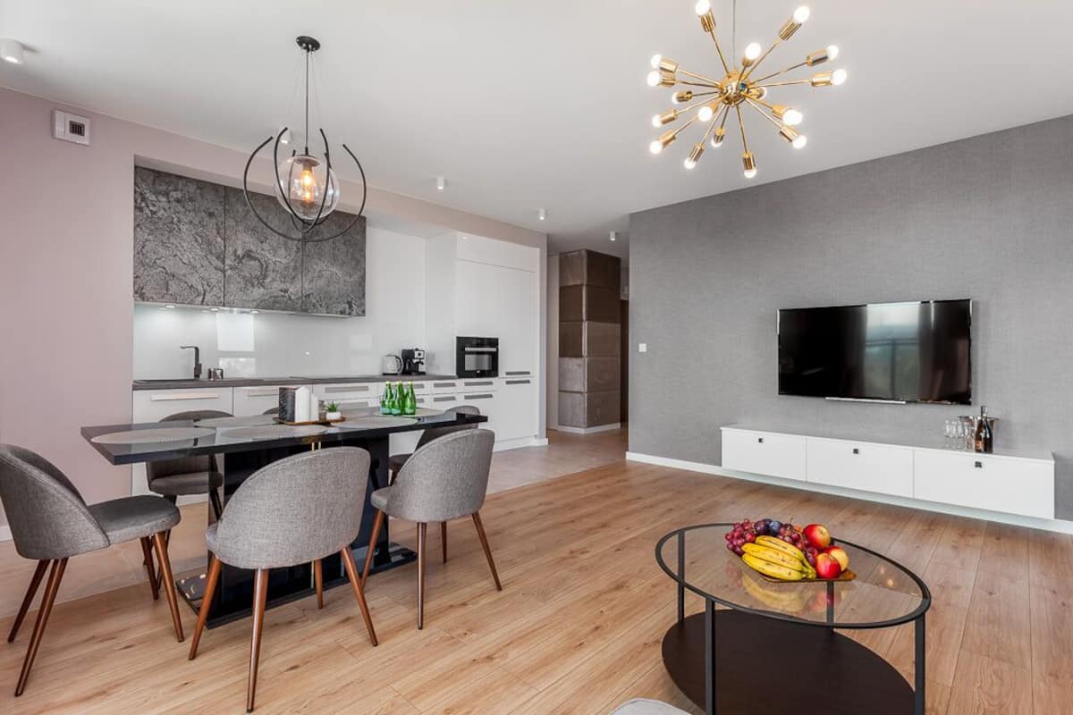 A modern kitchen with a central island, a dining area with chairs, a TV mounted on the wall, and a fruit bowl on the coffee table.