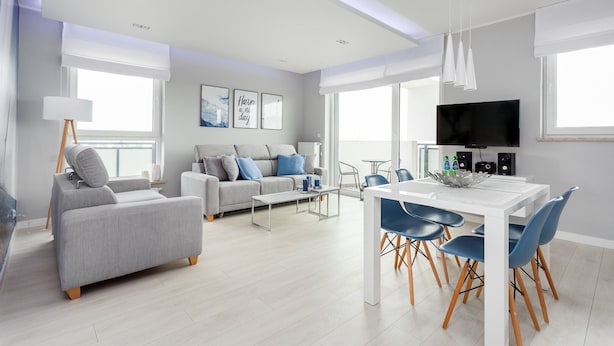 Ein modernes Wohnzimmer mit grauem Sofa, einem Essbereich mit blauen Stühlen und einem an der Wand befestigten Flachbildfernseher.
