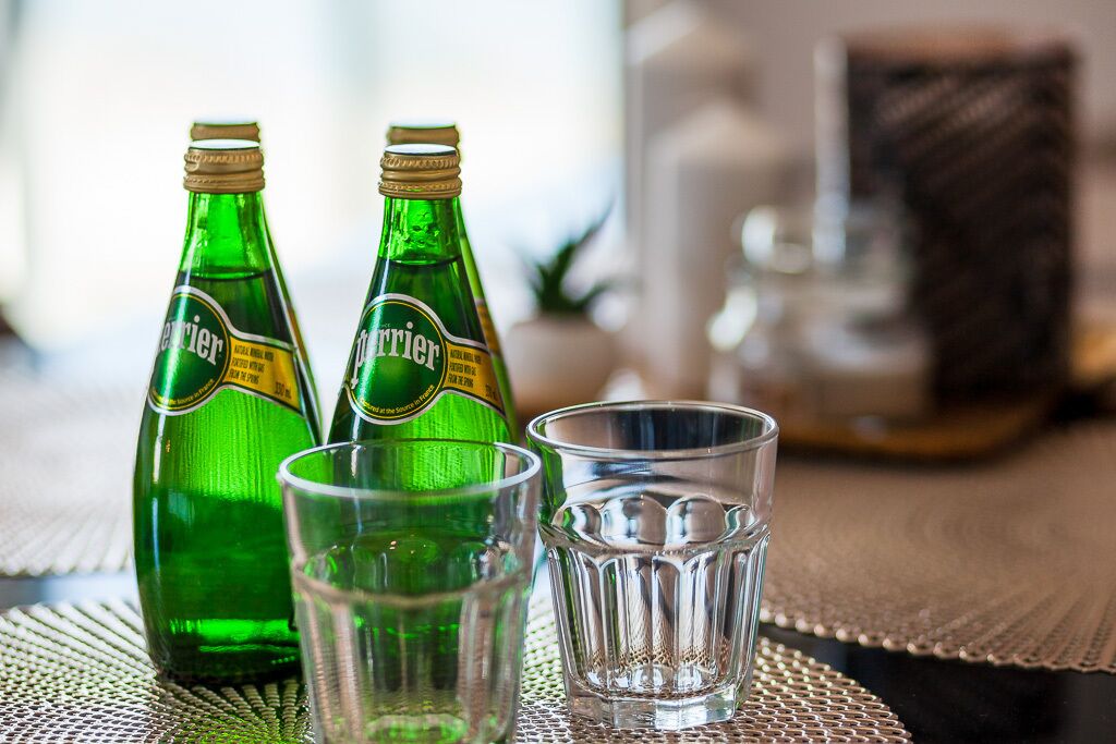Dos botellas verdes y dos vasos sobre una mesa con un posavasos tejido.