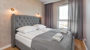 Ein ordentlich bezogenes Bett mit grauem Kopfteil und weißen Bettwäsche, ein Fenster mit Vorhängen sowie eine Wandleuchte.