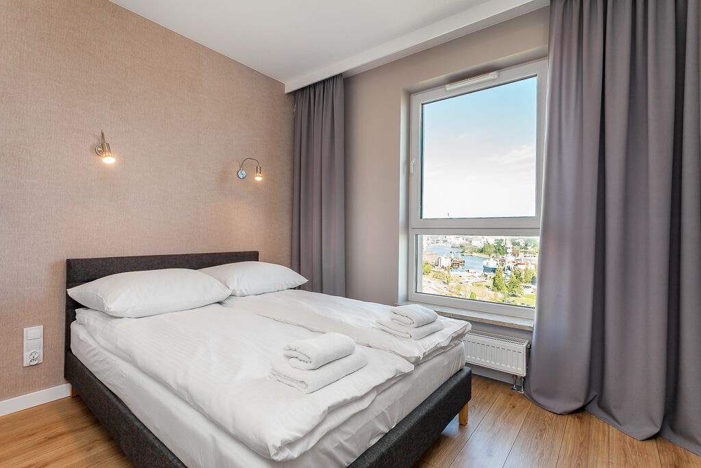 A neatly made double bed with white linens, a view of a cityscape through a large window, and a modern light fixture on the wall.