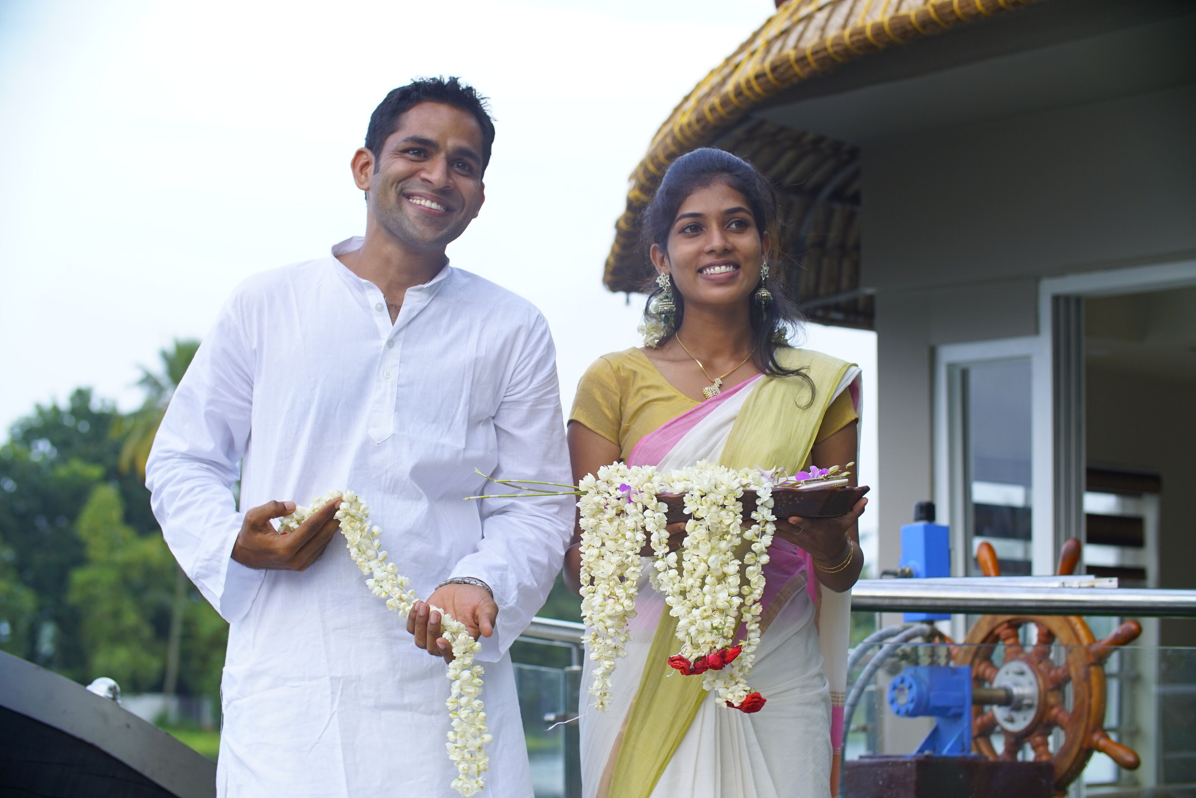 A man and a woman holding garlands, standing in front of a building with a visible steering wheel and a window.