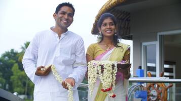 A man and a woman holding garlands, standing in front of a building with a visible steering wheel and a window.