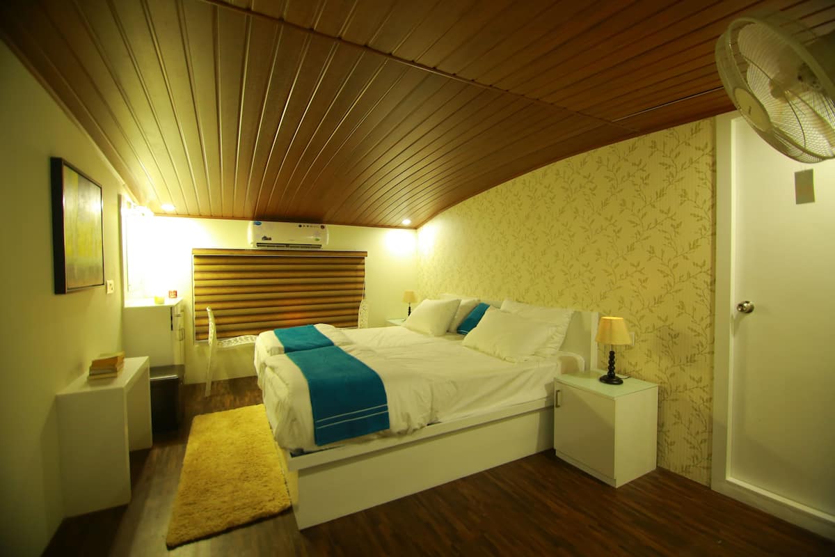 A bedroom with a wooden ceiling, a bed with a blue accent, a bedside table with a lamp, and a wall-mounted air conditioner.