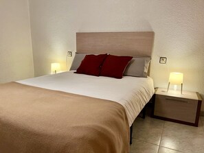 Un lit bien fait, avec une tête de lit beige, deux tables de chevet avec des lampes et un coussin rouge.