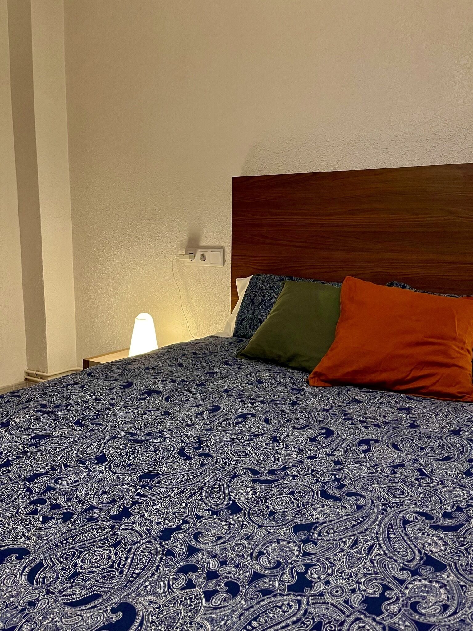 Ein ordentlich bezogenes Bett mit einem gemusterten Überwurf, einem Holz-Kopfteil und drei Kissen in verschiedenen Farben.