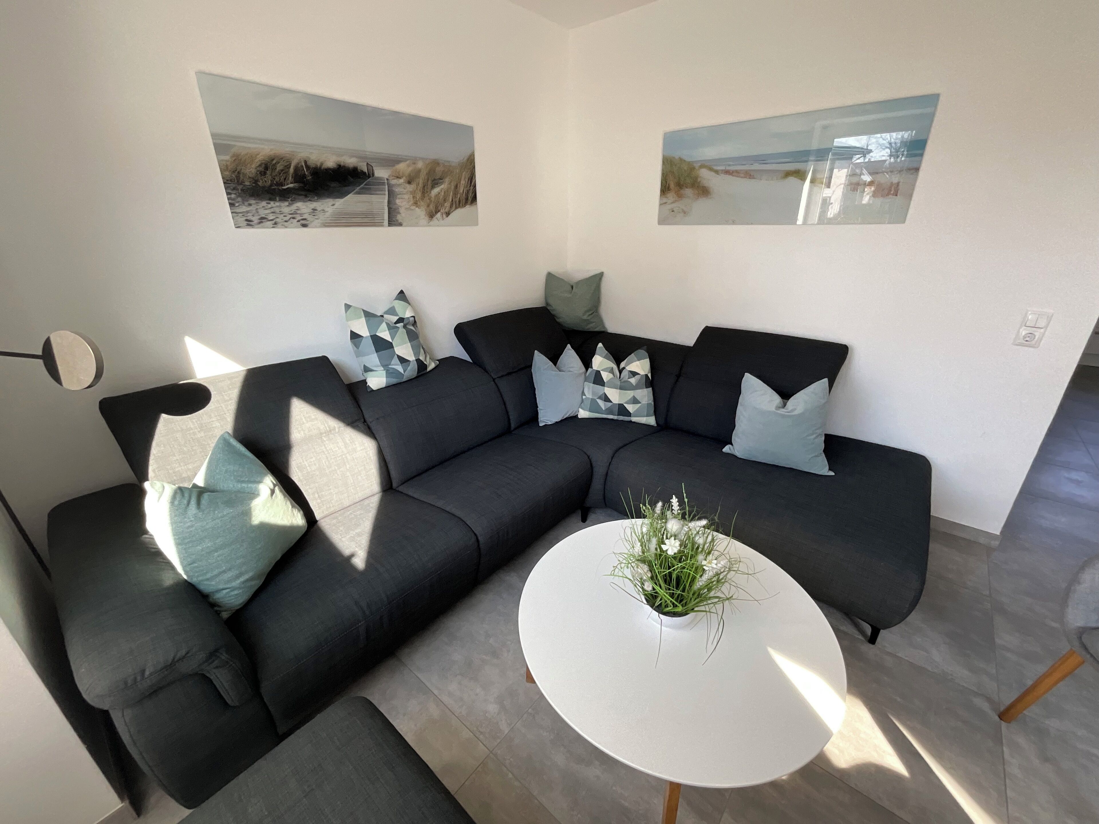 Ein modernes Wohnzimmer mit einem dunkelgrauen Ecksofa, einem runden weißen Couchtisch und zwei Wandbildern zum Thema Strand.