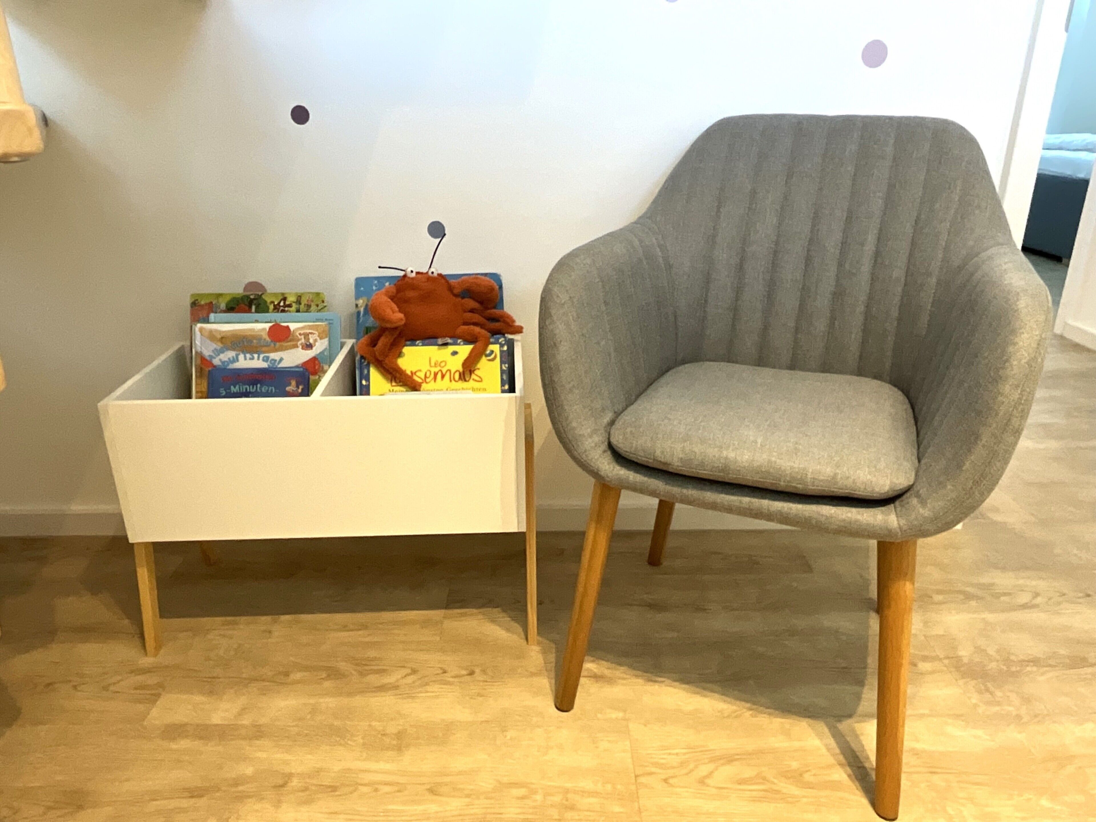 A room with a grey upholstered armchair and a white side table with books and a plush toy.