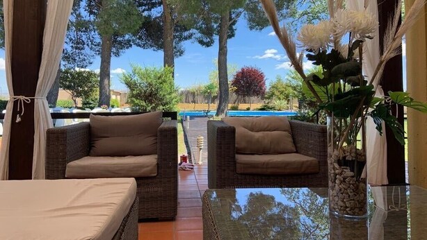 A covered outdoor seating area with wicker furniture and a glass table, overlooking a pool and trees. - Luxury villa with detail decorated pool (Zaragoza)