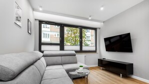A modern living room with a grey sectional sofa, a glass coffee table, a black TV stand, and a large window with blinds. - Frey Homes Superior Old Town (Gdansk)