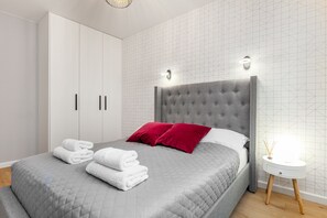 Ein ordentlich bezogenes Bett grau, mit roten Kissen, ein weißer Nachttisch mit Lampe und ein Kleiderschrank mit schwarzen Griffen.