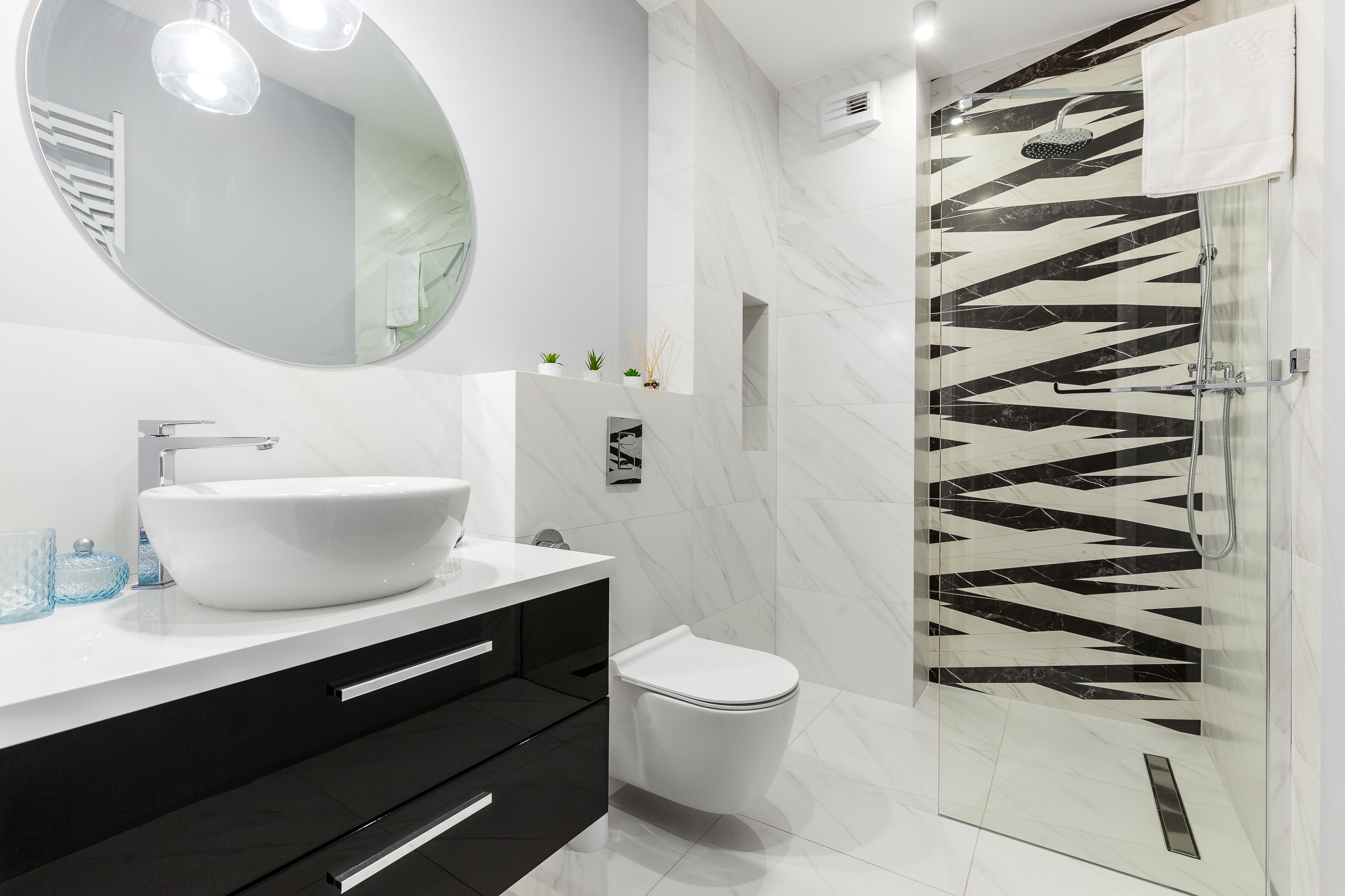 Casa de banho moderna com lavatório branco, armários pretos, sanita e cabine de duche em vidro com padrão de azulejos preto e branco.