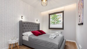 A modern bedroom with a grey bed, red pillows, a small bedside table, and a window with blinds. - Frey Homes Superior Old Town (Gdansk)