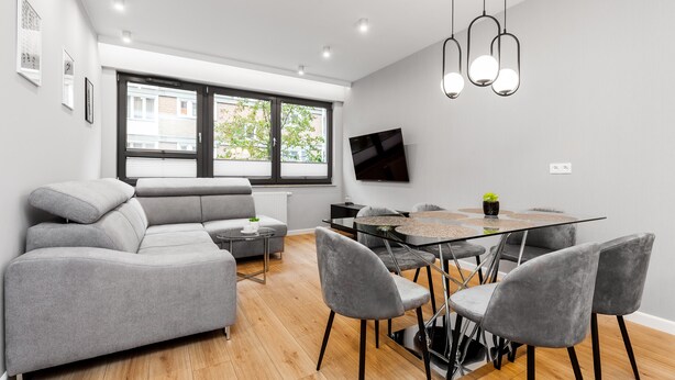A modern living room with a grey sofa, a glass dining table with grey chairs, a TV mounted on the wall, and a large window with blinds. - Frey Homes Superior Old Town (Gdansk)