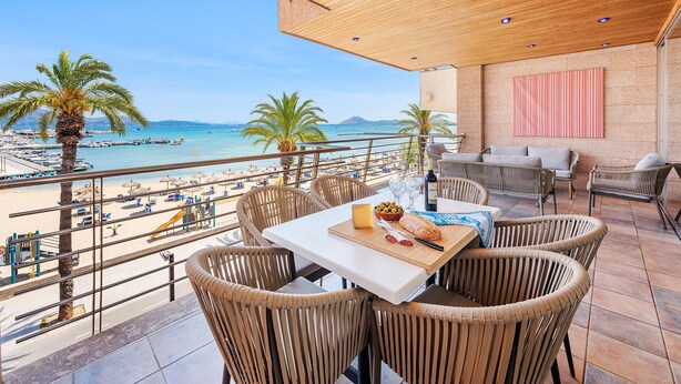 A terrace with wicker furniture, a table with food, and a view of the beach and sea. - Vistamar II frontline sea Apt. in a prime location in Puerto Pollensa (Puerto Pollensa)