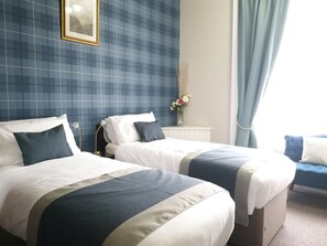 Une chambre d’hôtel avec deux lits, une table de chevet, un vase avec des fleurs et une fenêtre avec des rideaux.