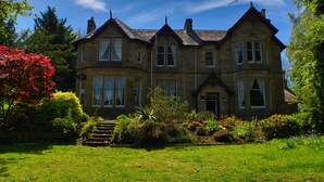 A large, stone building with multiple windows and a prominent entrance, surrounded by lush greenery and a well-maintained garden. - Heatherlie House Hotel (Selkirk)