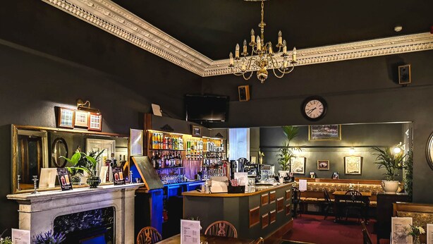 A bar area with a fireplace, bar counter, and seating. There are tables and chairs for dining, a chandelier, and a clock on the wall. - Heatherlie House Hotel (Selkirk)