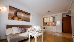 Een moderne woonkamer met een bank, een salontafel en een eethoek met stoelen. Er is een keuken met kastjes en een koelkast. Een groot schilderij hangt aan de muur.