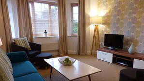 Un salon comprenant un canapé, une table basse avec un panier de fruits, un téléviseur posé sur un meuble et un mur orné d’un motif.