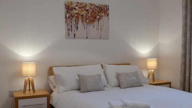 Un lit bien fait, avec du linge de lit blanc et des oreillers, deux lampes de chevet, une petite table de nuit en bois avec un tiroir et un tableau accroché au mur.