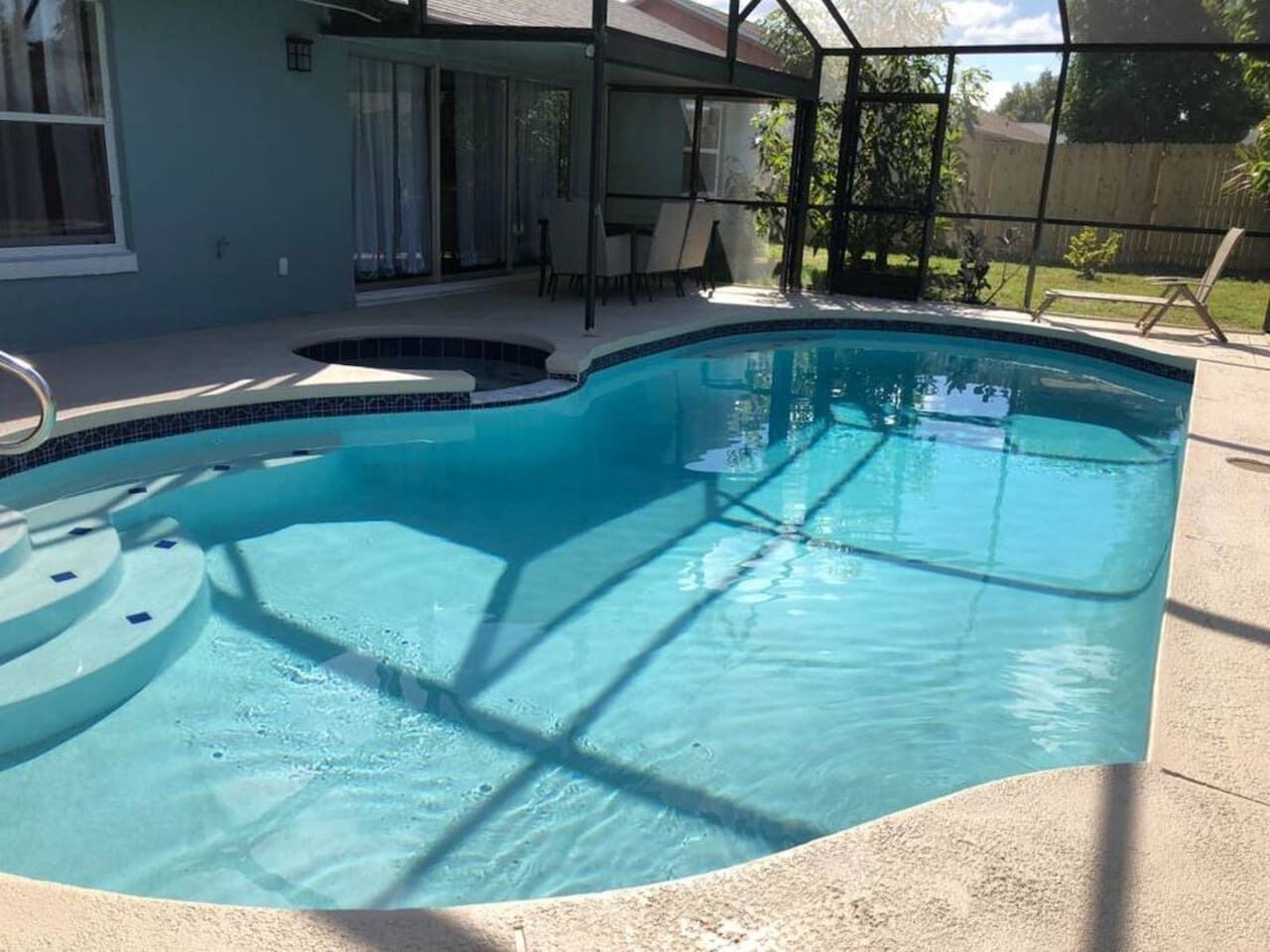 Ein Schwimmbad mit Sonnensegel, einer überdachten Terrasse und Blick auf einen Garten mit Zaun.