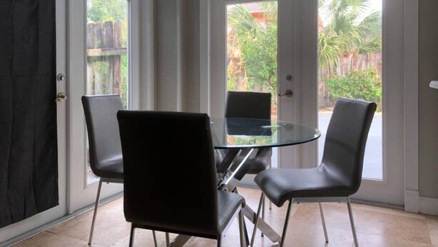 A dining area with a glass table and four chairs, large windows with a view of greenery, and a door with a black curtain. - 6151 Sandcrest · Next to Universal. Pool Sleep 9. Walk to Park (Orlando)