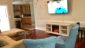 A living room with a TV, a fireplace, and a kitchen in the background. - 6151 Sandcrest · Next to Universal. Pool Sleep 9. Walk to Park (Orlando)