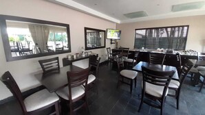 Un comedor con varias mesas y sillas, ventanas grandes con persianas y un televisor en la pared.