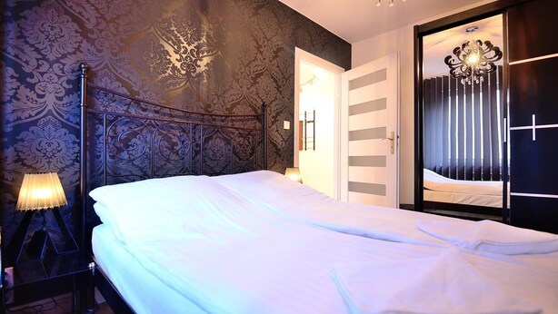 Een net opgemaakt bed in een kamer met behang met een patroon, een staande lamp en een kroonluchter.