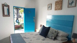 A bedroom with a blue headboard, a bed with a patterned bedspread, two pillows, a bedside table with a lamp, and a door leading outside. - Macaneta Holiday Resort (Marracuene)