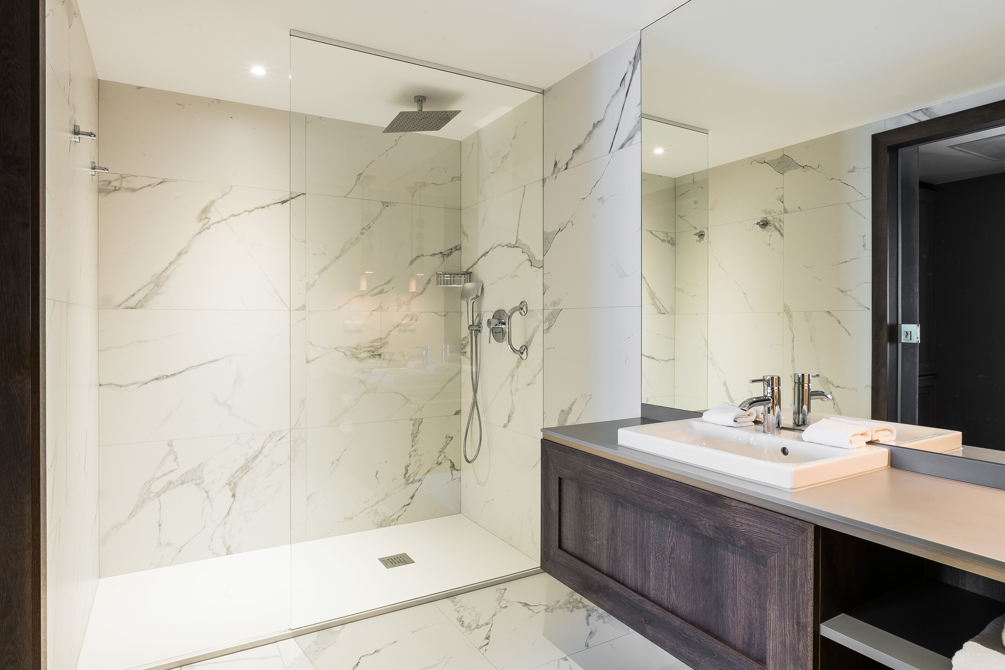 A modern bathroom with a glass-enclosed shower, marble walls, a dark wood vanity, and a large mirror.