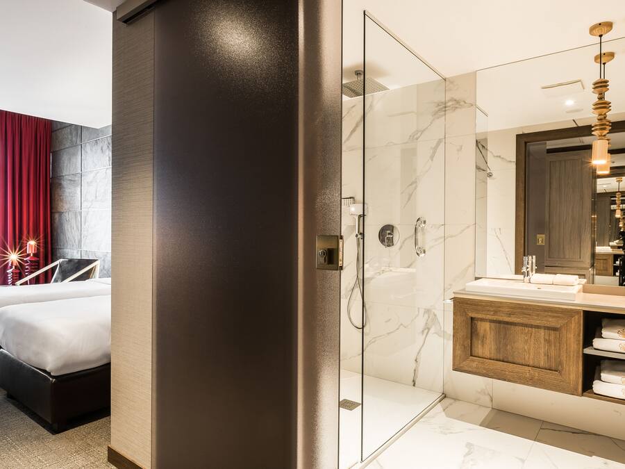 Een moderne hotelkamer met een douchecabine van glas, een houten wastafel met spiegel en een bed met een rood gordijn.