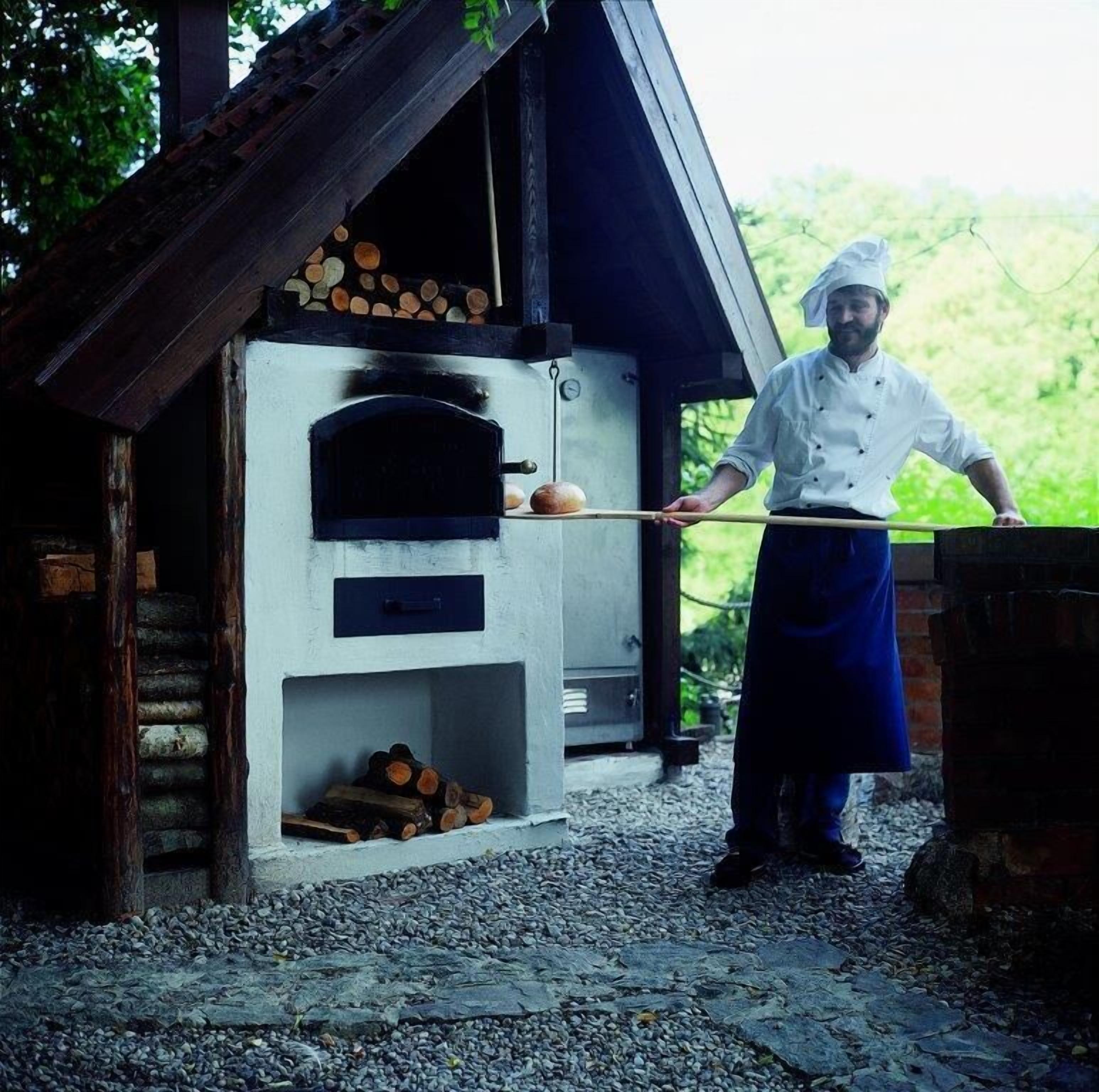 A chef in a white uniform and blue apron standing in front of a wood-fired oven with logs stored inside.