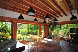 A spacious patio with multiple tables and chairs, large windows offering a view of a lush garden, and hanging pendant lights. - Młyn Klekotki spa resort (Gmina Godkowo)
