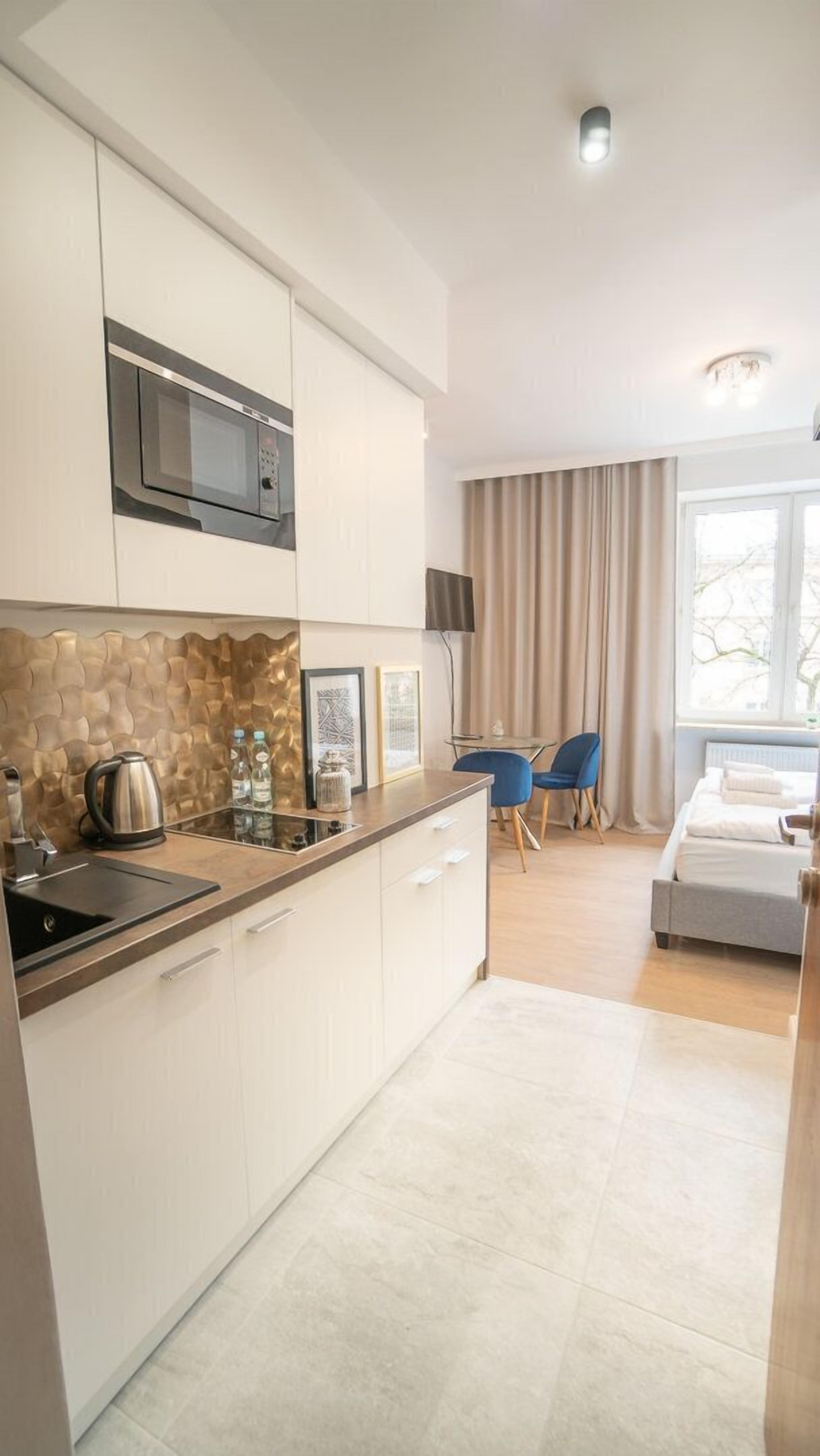 Eine moderne Küche mit Mikrowelle, Wasserkocher und Wasserflaschen auf der Arbeitsfläche. Es gibt einen Essbereich mit Tisch und Stühlen, und ein Bett im Hintergrund.