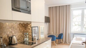 A modern kitchen with a microwave, kettle, and water bottles on the counter. There is a dining area with a table and chairs, and a bed in the background. - Frey Homes City Centre Plac Zbawiciela (Warsaw)