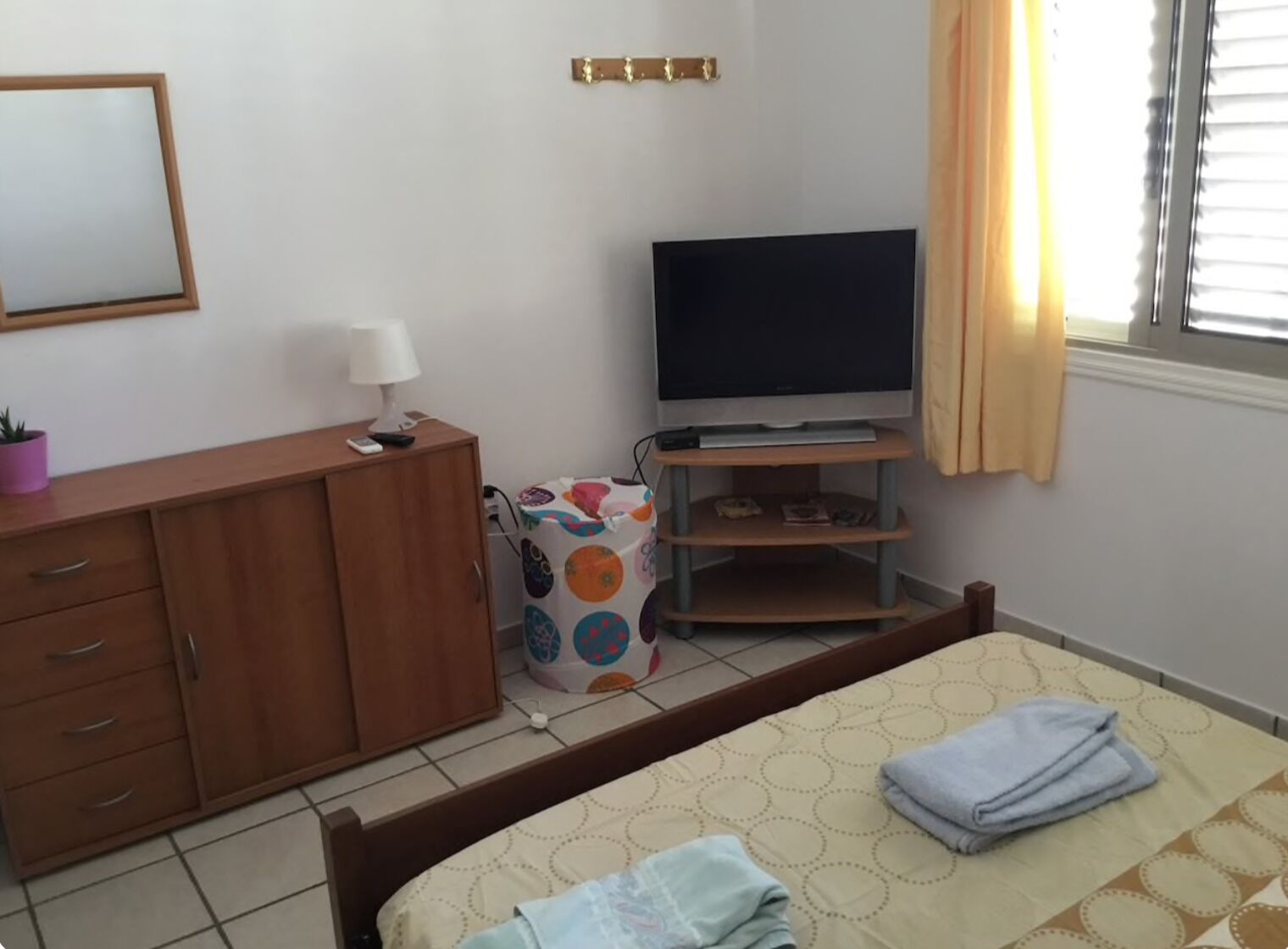 Une chambre à coucher avec un lit, un téléviseur posé sur un meuble, une armoire, une table de chevet avec une lampe et une fenêtre avec des rideaux.