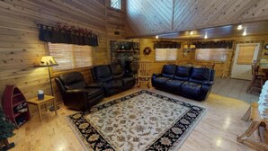 A cozy cabin interior with wooden walls, a fireplace, two leather sofas, a dining area, and a small table with a lamp.