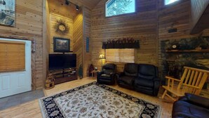 A cozy log cabin interior with a fireplace, leather sofas, a TV, and a large rug.