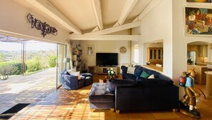 A spacious living room with a large sofa, a television, and a bookshelf. There is a balcony with a view of the countryside. - Charming 5 bedroom, 3 bathroom Villa in gated domain near Valbonne with sea view (Opio)