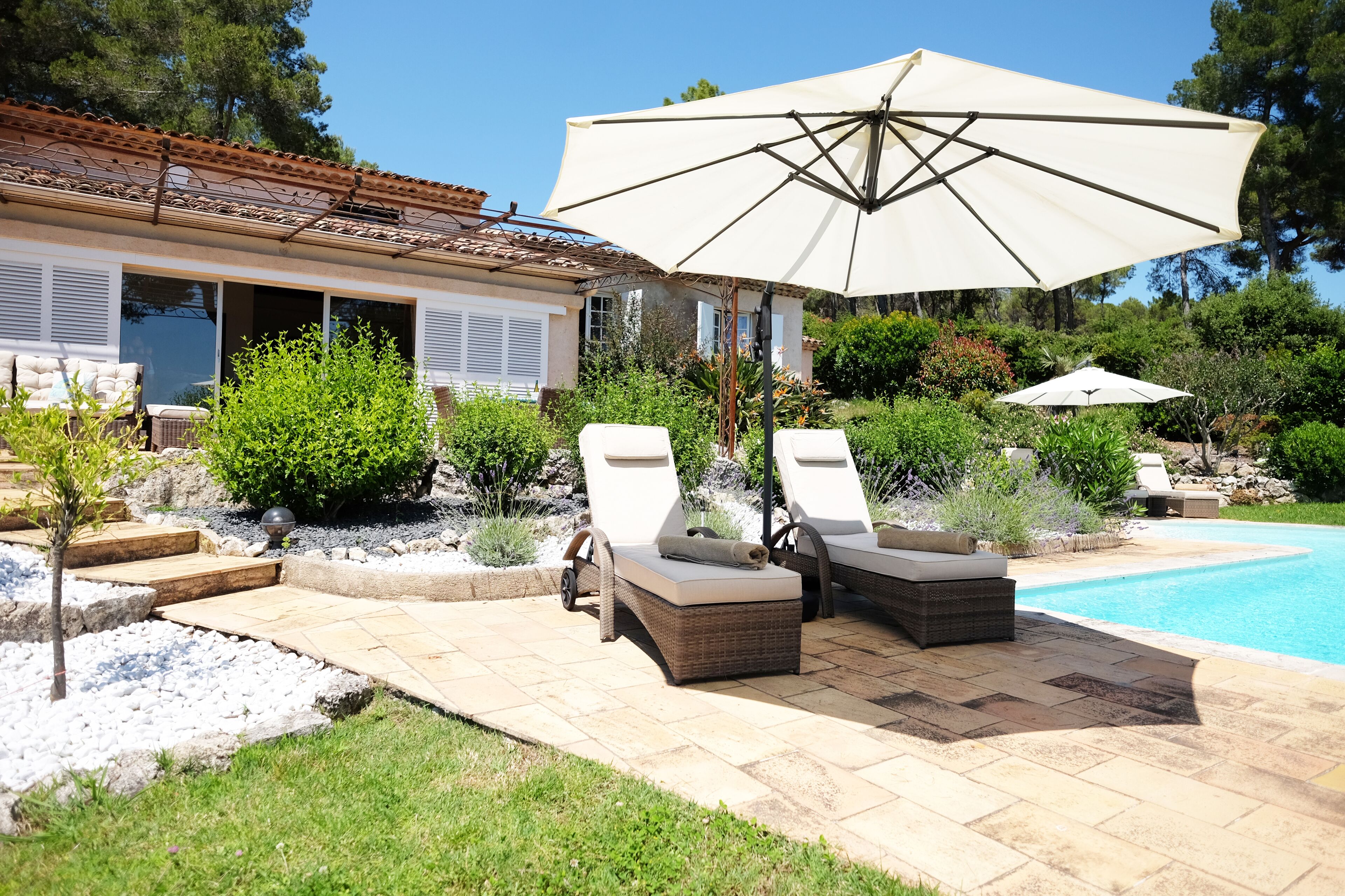 Zona de piscina amb dues gandules i un gran parasol, envoltada d'un jardí ben cuidat i una casa amb teulada de rajola.