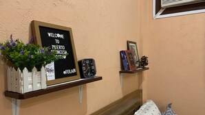 Una habitación con una cama, un cartel de bienvenida, un reloj, una estantería con libros y una maqueta, y una ventana con persianas.