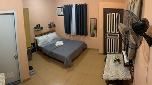 Un dormitorio con cama, mesita de noche, espejo, aire acondicionado de pared y ventilador de techo, junto a una pequeña mesa con un florero.