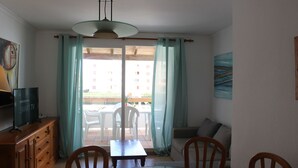 A dining area with a glass table, wooden chairs, a sofa, and a TV on a wooden cabinet. - Deltamar 24 Solo Familias! (Xeraco Playa)